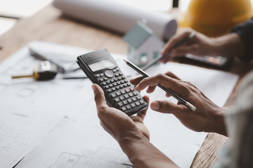 Person using a calculator over building plans while considering the question is roofing business profitable.